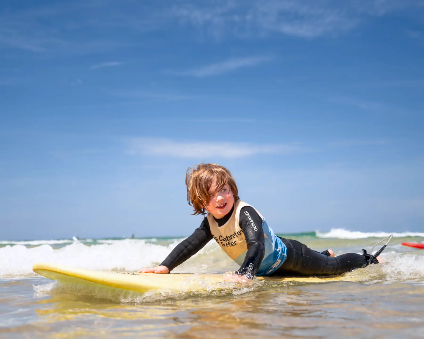 A child lying on a yellow surfboard, smiling while learning to surf in shallow waters, illustrating kids' surf lessons