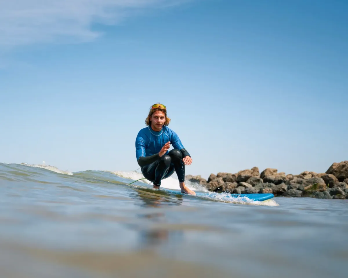 A surfer in a blue wetsuit sitting on a surfboard in calm waters, with a clear blue sky and rocky shoreline in the background, showcasing ideal surfing conditions