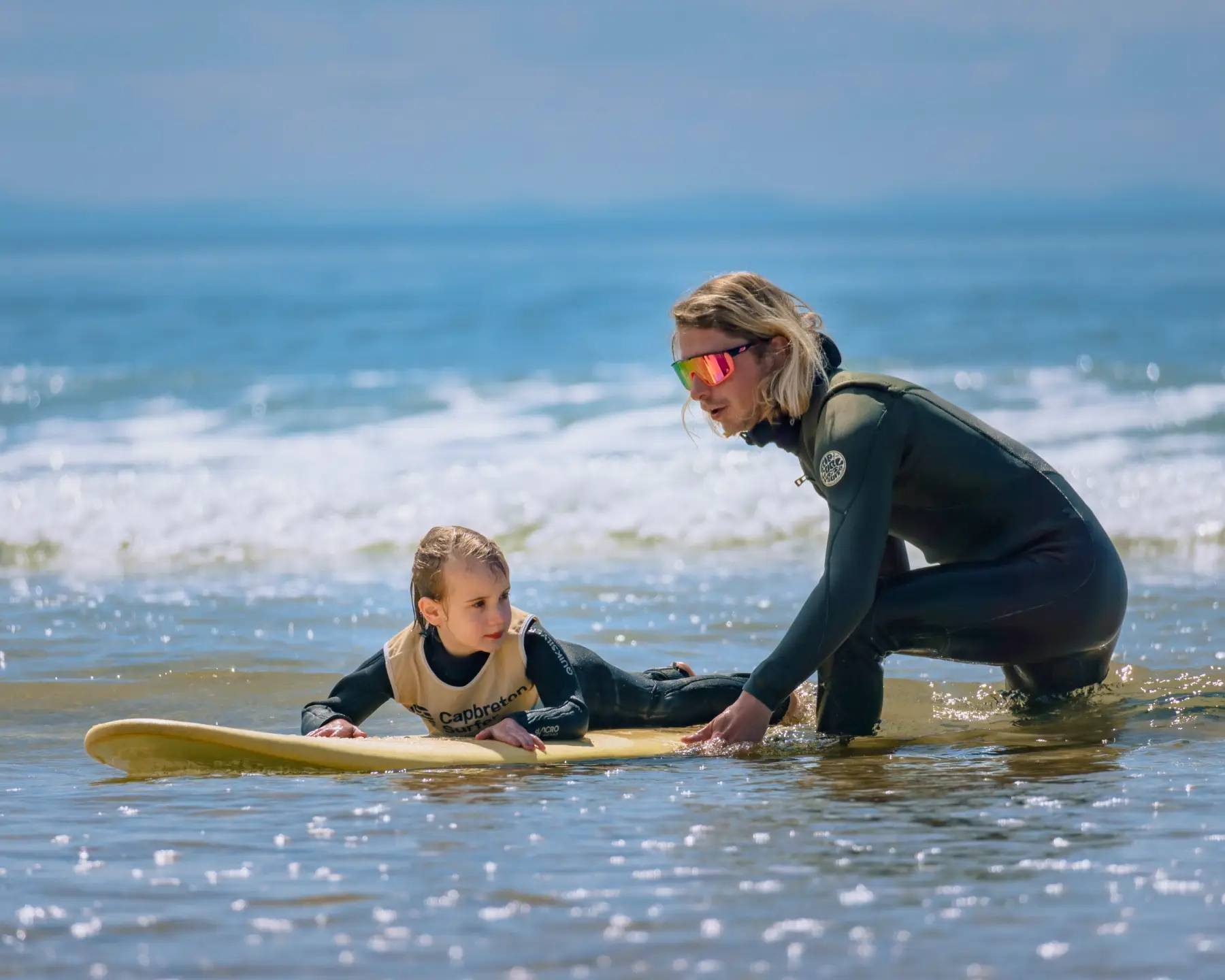 An instructor assisting a young girl on a surfboard in shallow waters