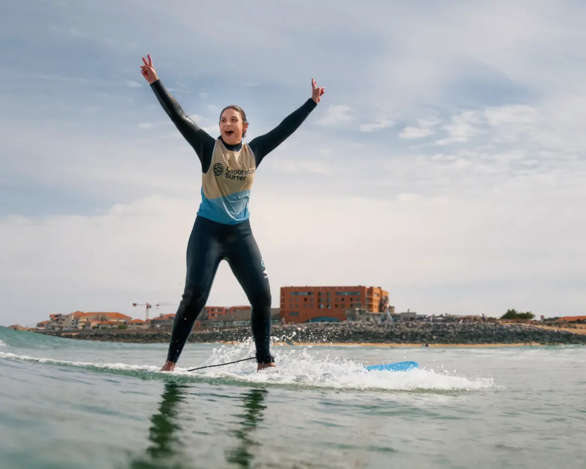 A smiling surfer in a black wetsuit with a beige jetsey, raising her arms in victory on her surfboard after managing to ride a wave standing up without falling off the board, illustrating the joy of surf lessons