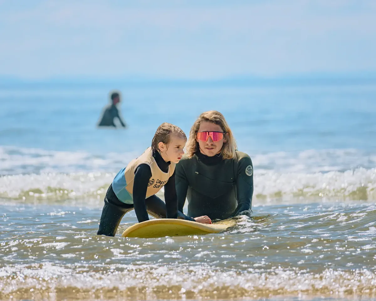 A surf instructor in a black wetsuit talking to a young girl and helping her climb on her surfboard in shallow waters