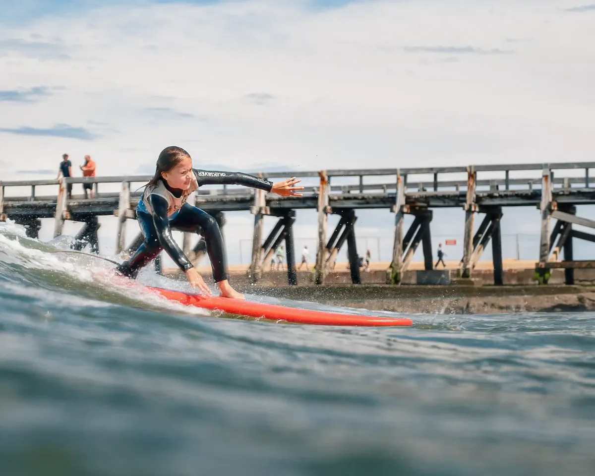 A young surfer in a black wetsuit, standing on a red surfboard, riding a wave with a pier in the background