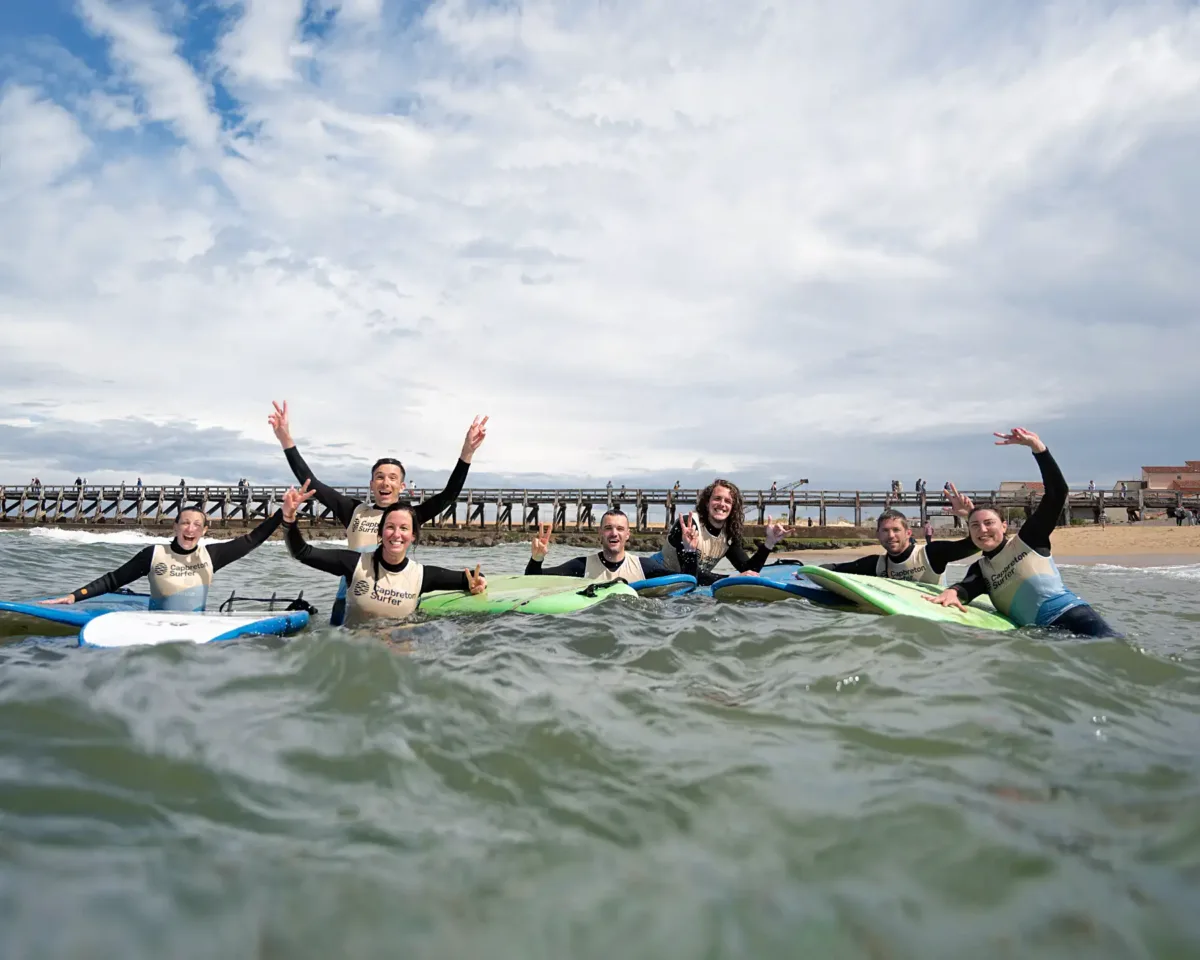 A group of adults in wetsuits stand in the water with surfboards, smiling and raising their hands in the air while making “V” signs with their fingers.