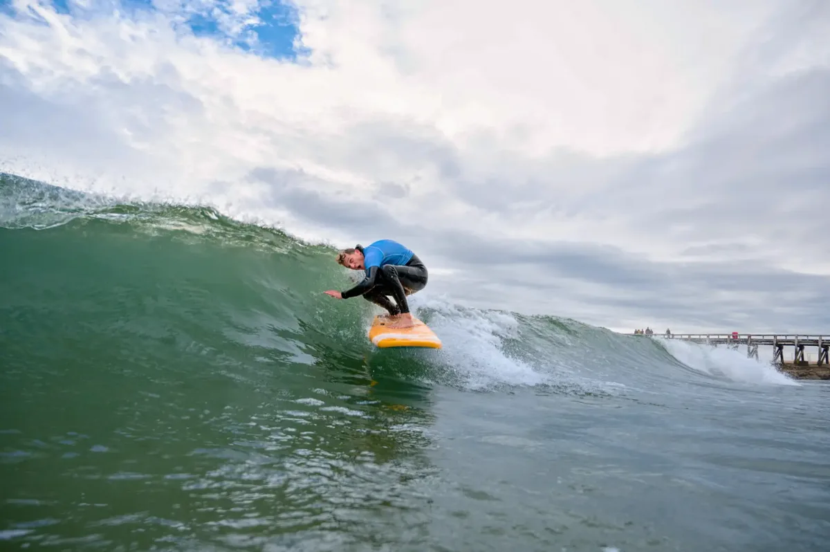 A surf instructor in black wetsuit and blue lycra rides a wave that's about to break, jesting and striking an incongruous pose.