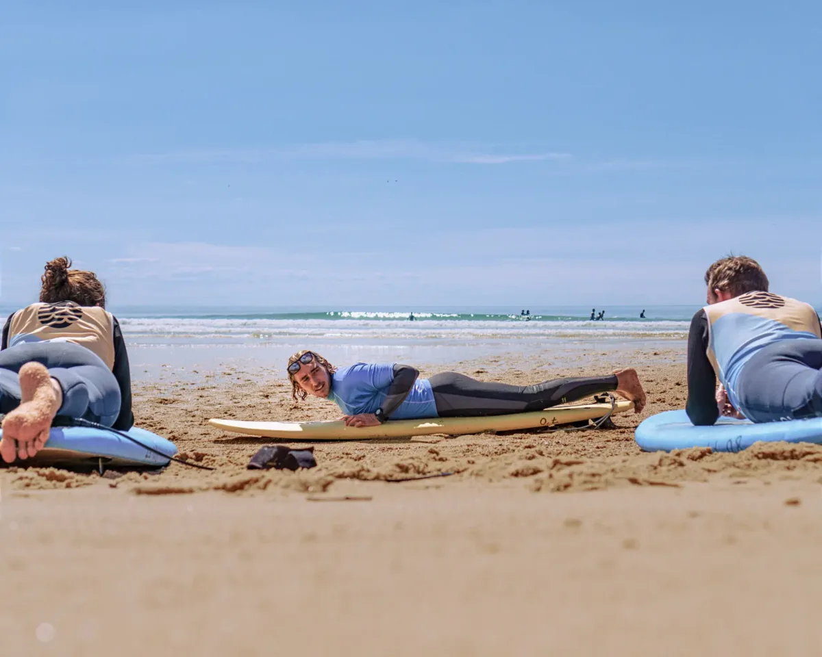A surf instructor, lying on his board on the beach, demonstrates techniques to two adults who are watching him while also lying on their boards. In the background, the ocean and waves can be seen in the distance, under a clear blue sky.