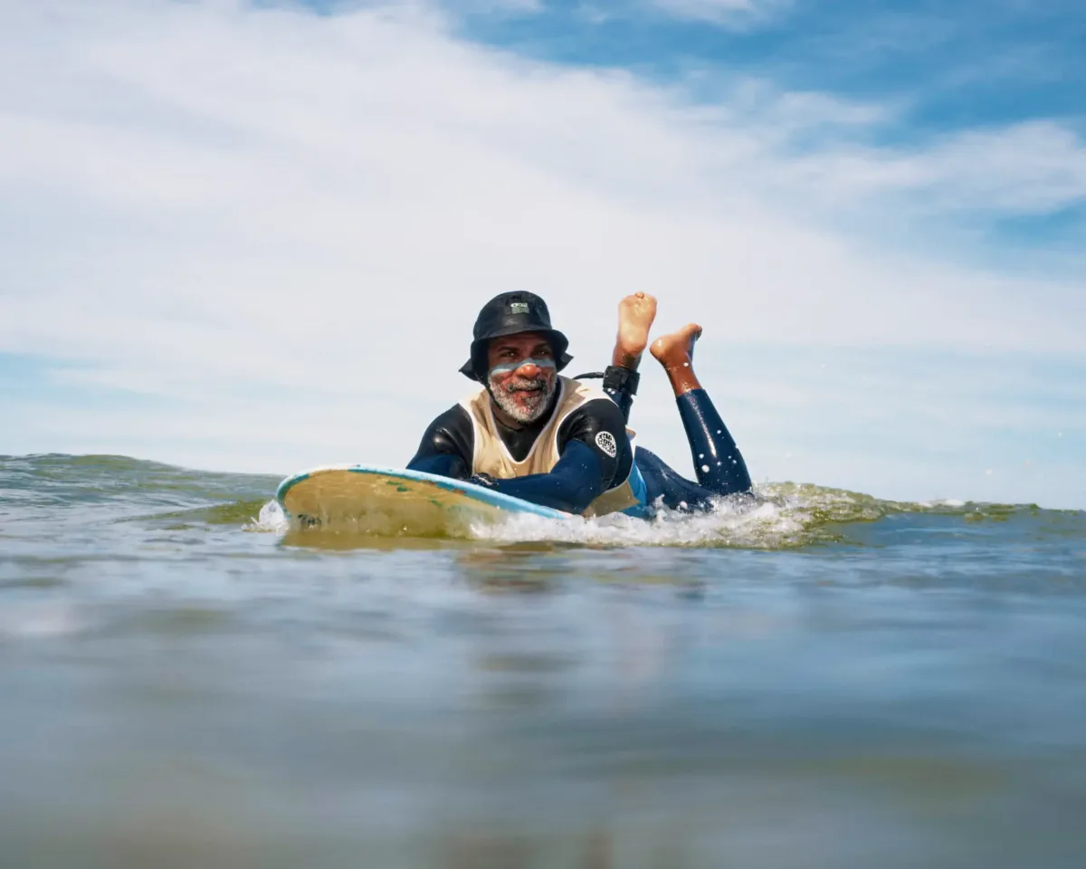A man wearing a black hat and wetsuit lies on a surfboard in the ocean, smiling and looking towards the camera, with sky and clouds in the background.