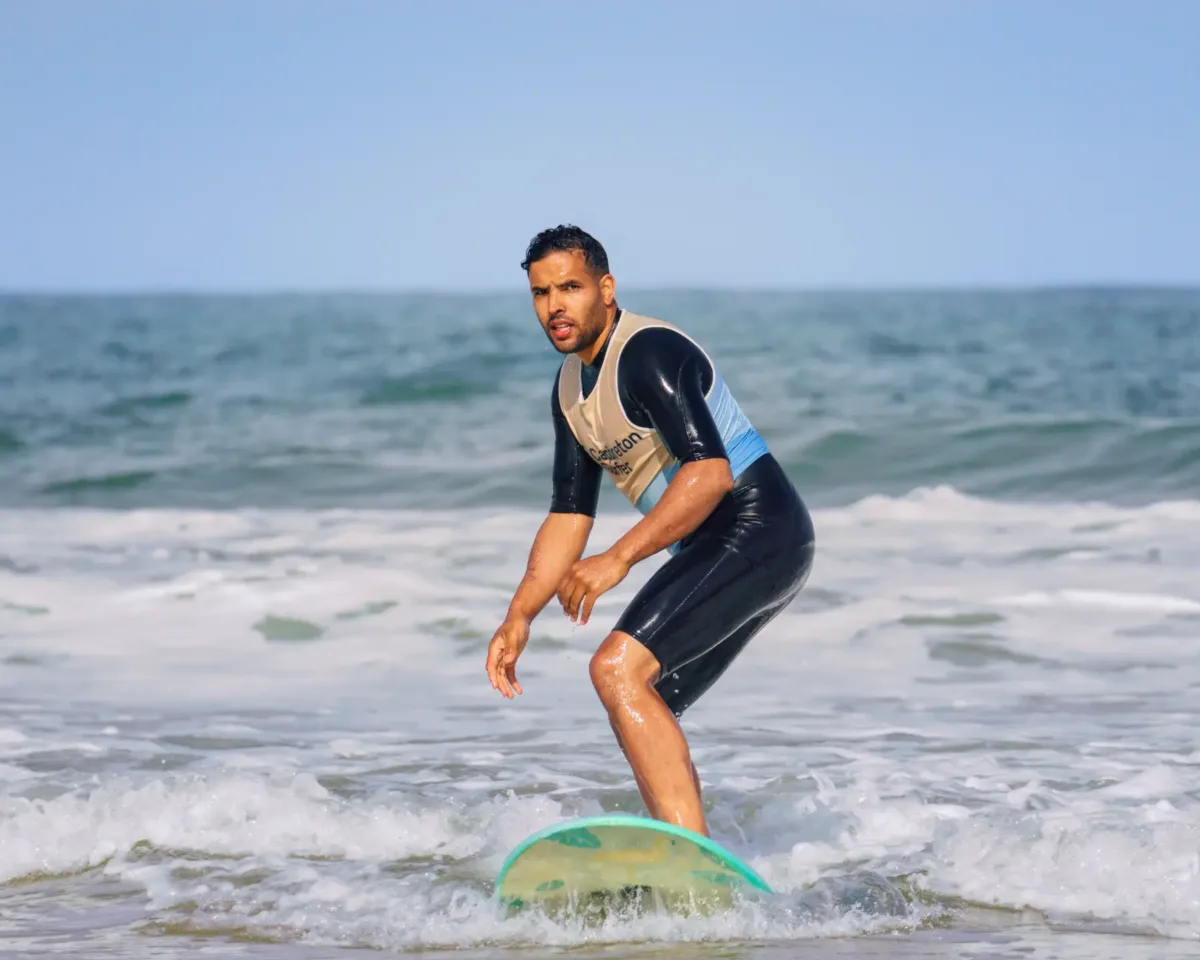 A man in a wetsuit faces the camera while standing on a surfboard in the ocean, with small waves around him.