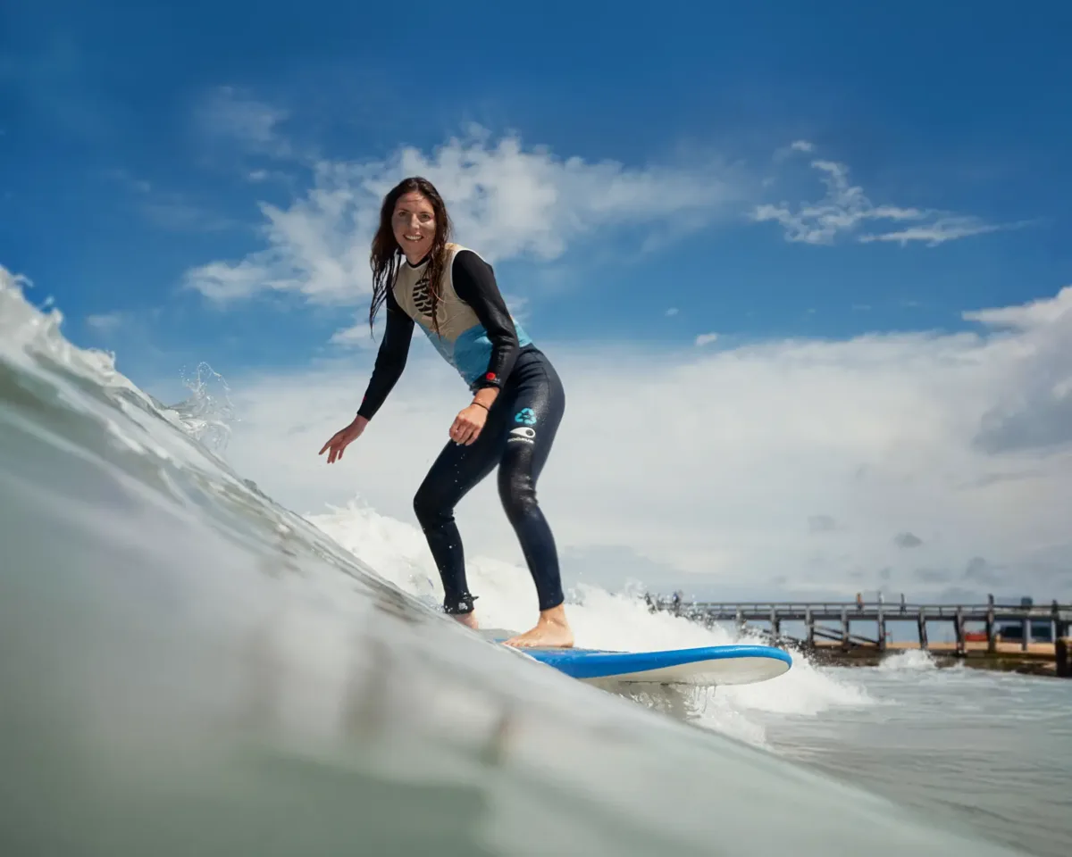 A woman wearing a wetsuit rides a wave on a blue surfboard towards the camera, under a partly cloudy sky.