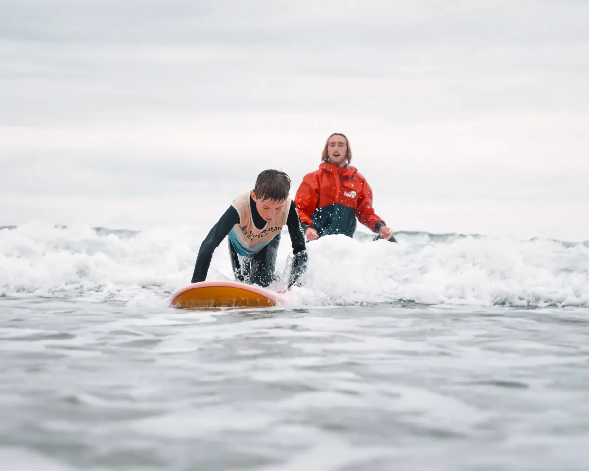 A kid on all fours on his surfing board, in the foam, in shallow waters, being closely watched by an instructor in a red coat who is in the water as well
