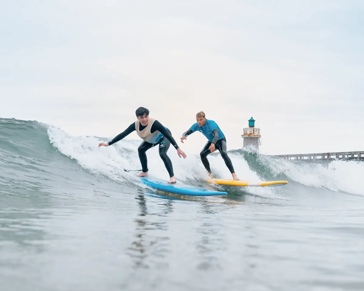 A teenager surfs a wave standing up on his blue surfboard, while the surf instructor, also surfing the same wave alongside him, gives him advice on his posture.