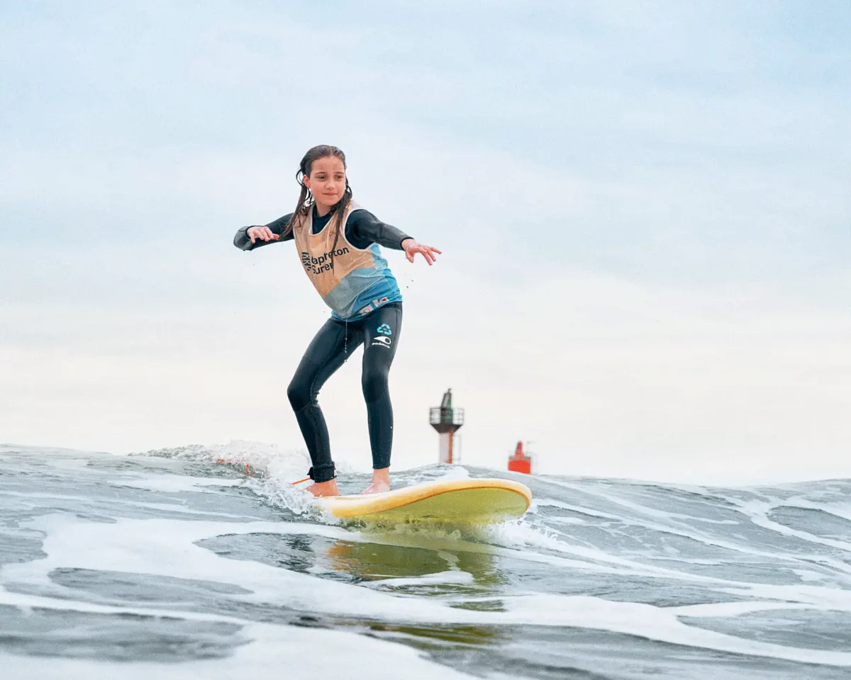 A teenager wearing a wetsuit and beige and blue lycra rides a yellow surfboard on a calm wave, with the Capbreton lighthouse visible in the distance.