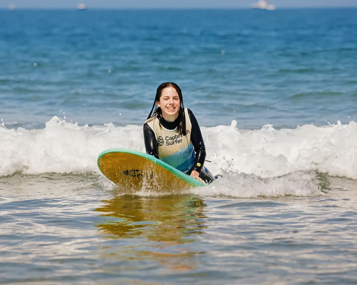 A teenager in a wetsuit and surf school vest lays on a surfboard in calm ocean waters with waves in the background. She is smiling.