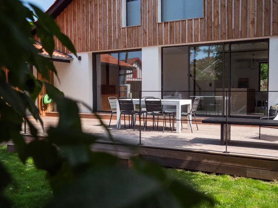 Wooden terrace with a table and chairs in front of the house’s large sliding glass doors.