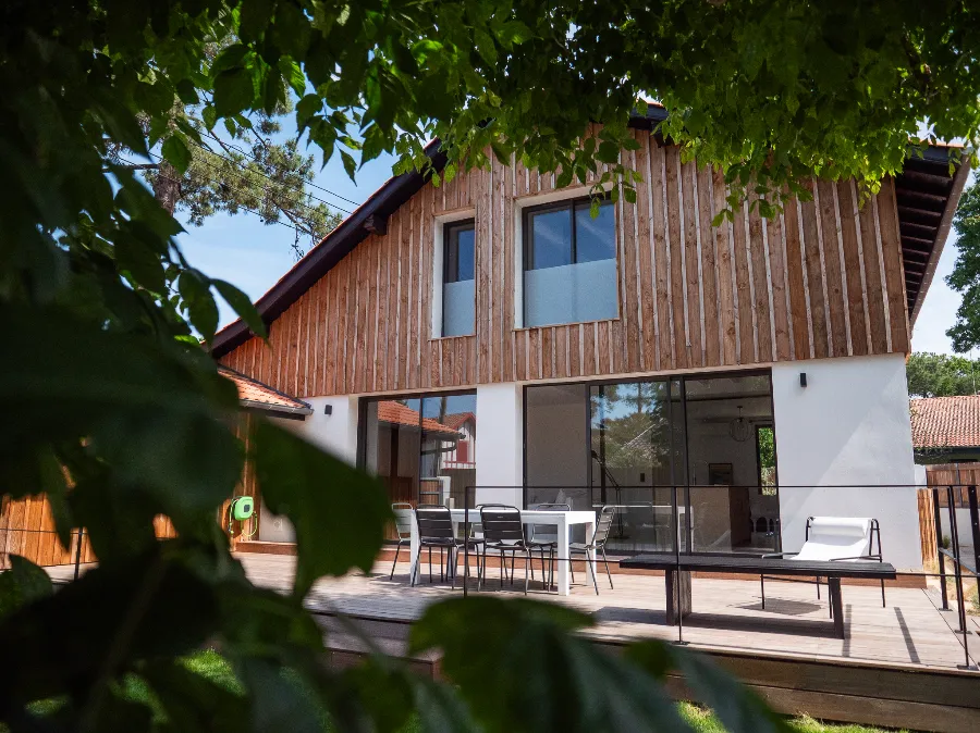 Wide view of the timber-clad house and its terrace, framed by foliage in the garden.