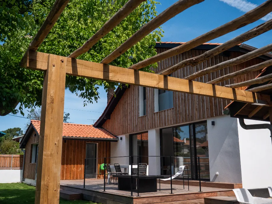 Timber-clad house viewed from the pergola, with a terrace, outdoor furniture, and lawn.